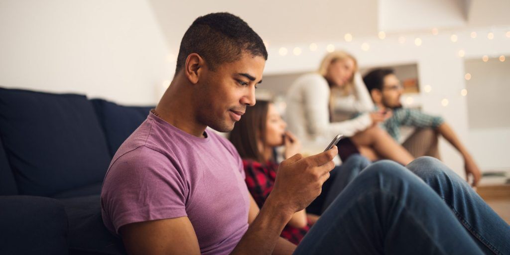 A man is sitting on a couch looking at his phone, likely awaiting lab testing results. In the background, three other people are sitting and talking quietly.