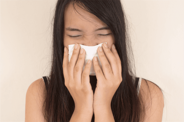 A person with long dark hair is sneezing or blowing their nose into a tissue, covering their nose and mouth with both hands at an urgent care.