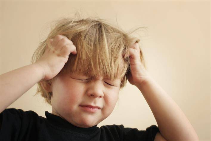 A young child with closed eyes and a frustrated expression pulls at their hair with both hands, standing outside the bustling medical center.