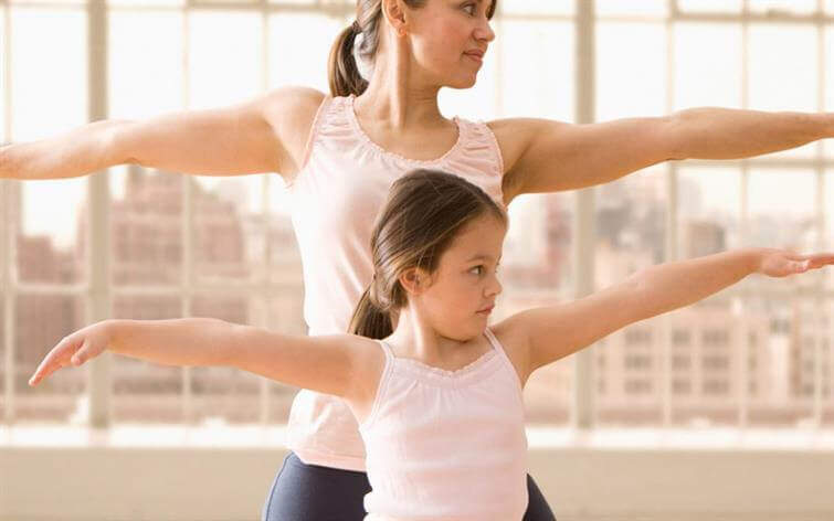 A woman and a girl perform a yoga pose together in front of large windows with a cityscape in the background, reflecting a serene contrast to the nearby medical center. Both are wearing light pink sleeveless tops.