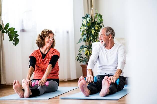 Two elderly individuals sitting on yoga mats in a bright room with plants, smiling and stretching with small dumbbells in their hands, appear rejuvenated after their visit to the nearby medical center.