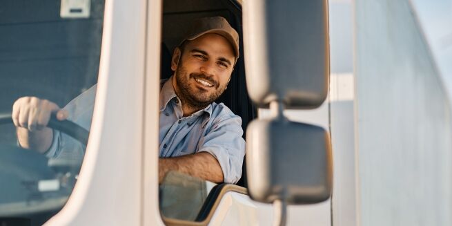 A man wearing a cap sits in the driver's seat of a vehicle, smiling and looking out the window, possibly on his way to an appointment at an American Family Care center.