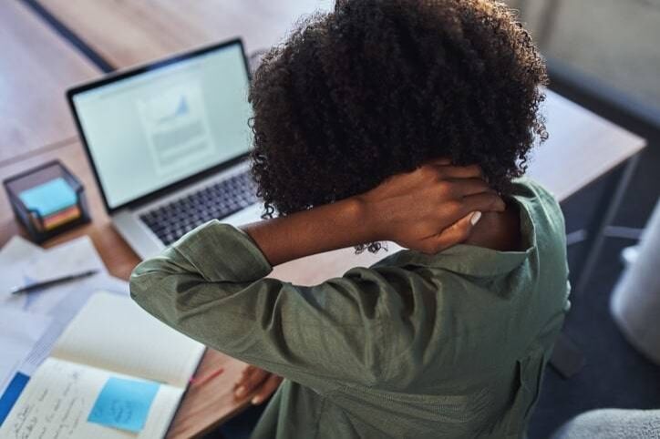 Person in green shirt sitting at a desk, rubbing their neck while looking at an open laptop with documents scattered around, possibly considering a visit to urgent care.