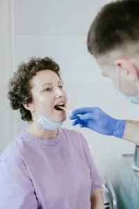 A person wearing a lavender shirt and a half-lowered face mask is having their mouth swabbed by a gloved healthcare worker at an urgent care clinic.