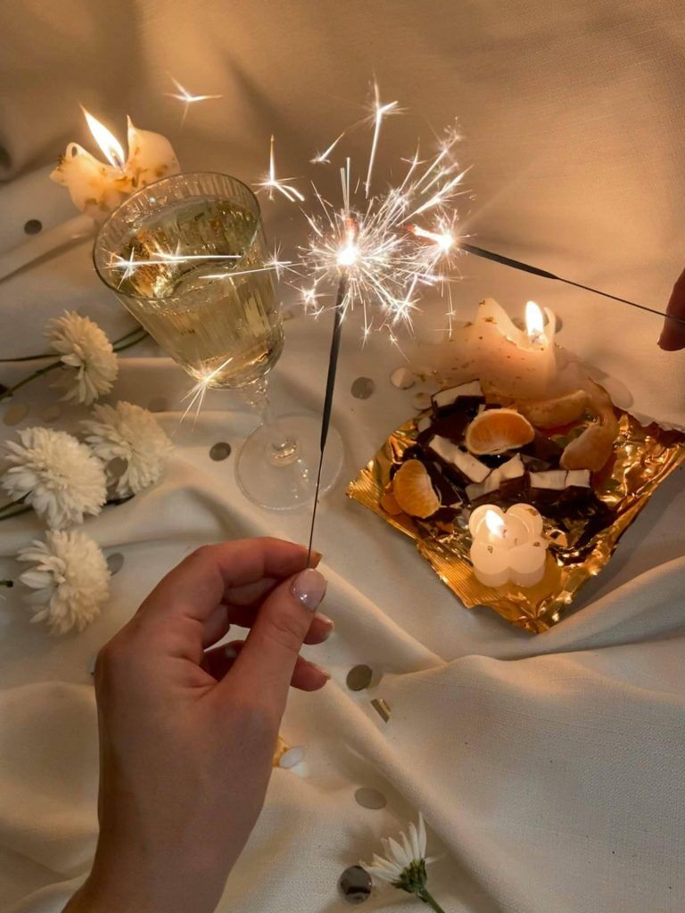 A person holding a lit sparkler near a glass of champagne, candles, white flowers, and a plate with orange slices and chocolate pieces on a white cloth looks like the perfect celebration setting before heading to an evening at the urgent care medical center.