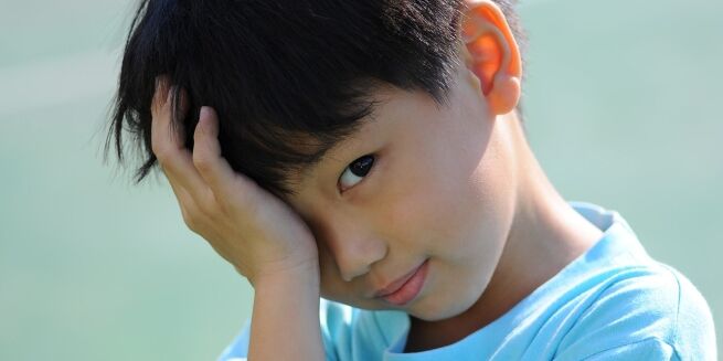 A child with short dark hair wearing a light blue shirt holds their hand to their forehead, gazing directly at the camera with a slight smile, perhaps waiting at the medical center for a routine physical.