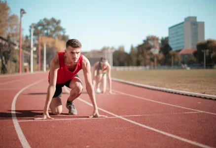 Two athletes in red gear are at the starting line of an outdoor track, preparing for a race, with urgent care professionals standing by to ensure their safety.