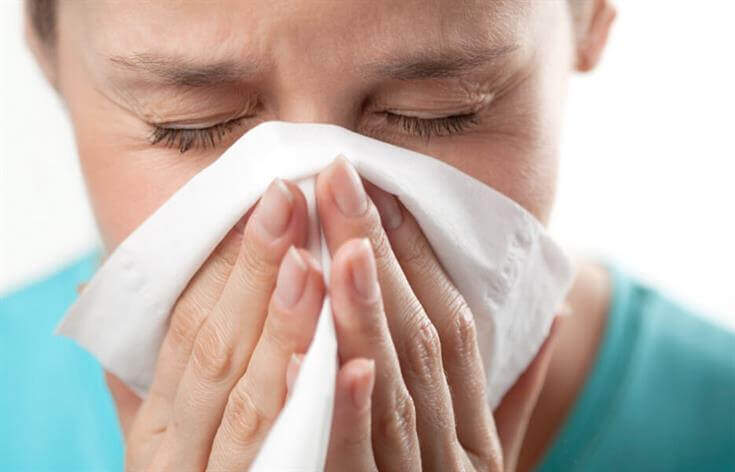 Person in a teal shirt sneezing or blowing their nose into a white tissue while at an urgent care clinic.