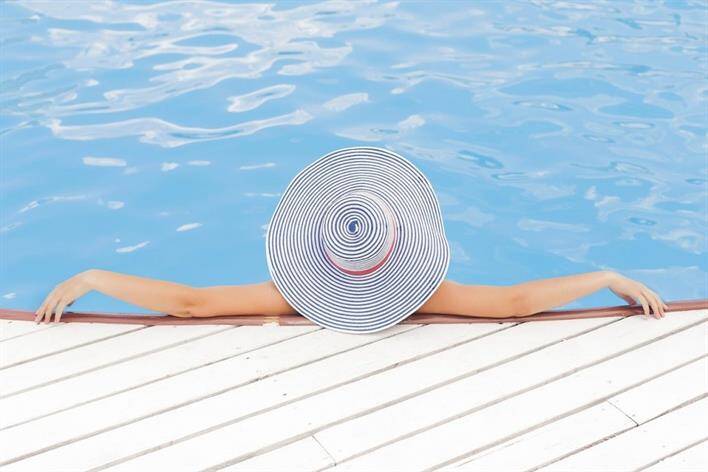Person wearing a large striped sun hat relaxing in a swimming pool, leaning on the pool's edge with arms outstretched. Nearby, the signage for an urgent care center hints at modern convenience blending seamlessly with leisure.
