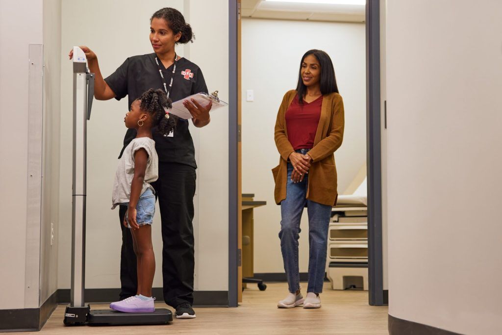 A healthcare worker measures a young girl's height while holding a clipboard at a medical center. A woman stands nearby in the hallway, observing the process.