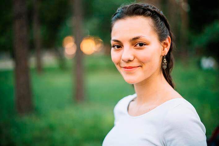 A young woman with braided hair smiles slightly while standing outdoors. Trees and greenery form the blurred background. She wears a white shirt and earrings, perhaps after a visit to an urgent care clinic for a routine check-up.